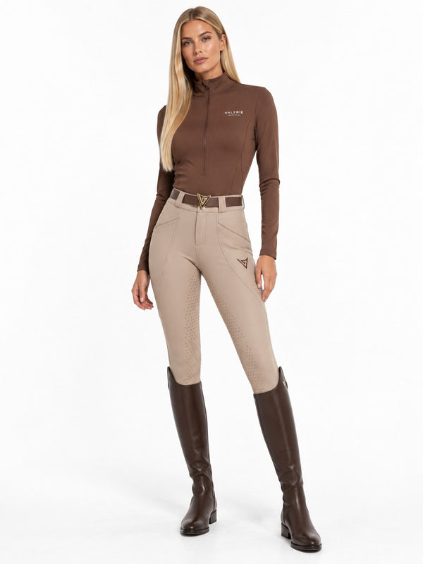 Rider in beige competition breeches with a chocolate zip-up turtleneck and brown tall boots, full-length front view — Valerie Equestrian Impulsion